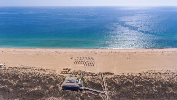 An aerial view of a beach with a clear sky, a sandy shore, and a small building with a dock. - Stunning beach house, panoramic views across Meia Praia bay (Lagos)