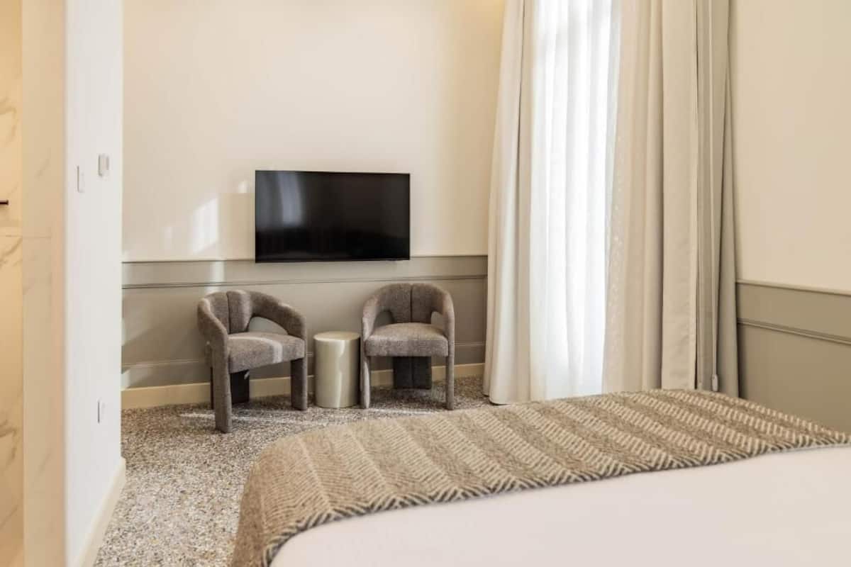 A hotel room with a bed, two armchairs, a small table, a flat-screen TV mounted on the wall, and a window with curtains.