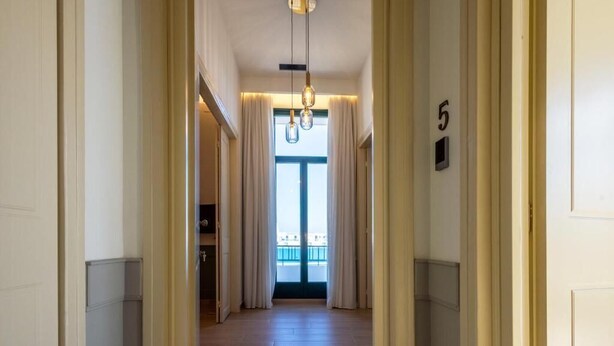 Un couloir d’hôtel avec le numéro « 5 » affiché sur le mur, un luminaire au plafond et une vue sur un balcon à travers une porte.