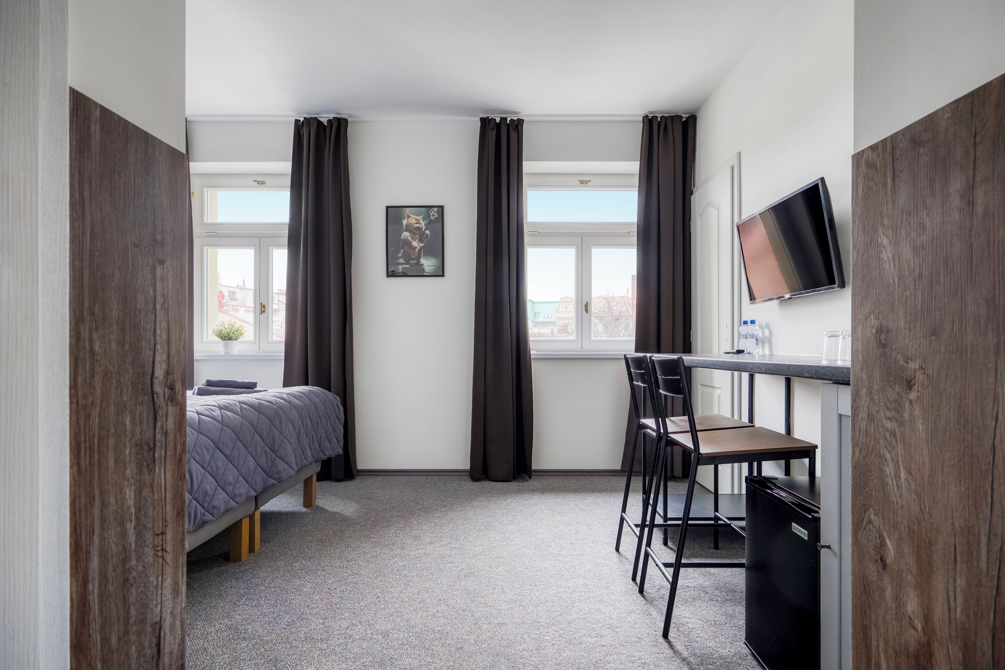 A compact room with a bed, a desk with chairs, a television, and a window with curtains.