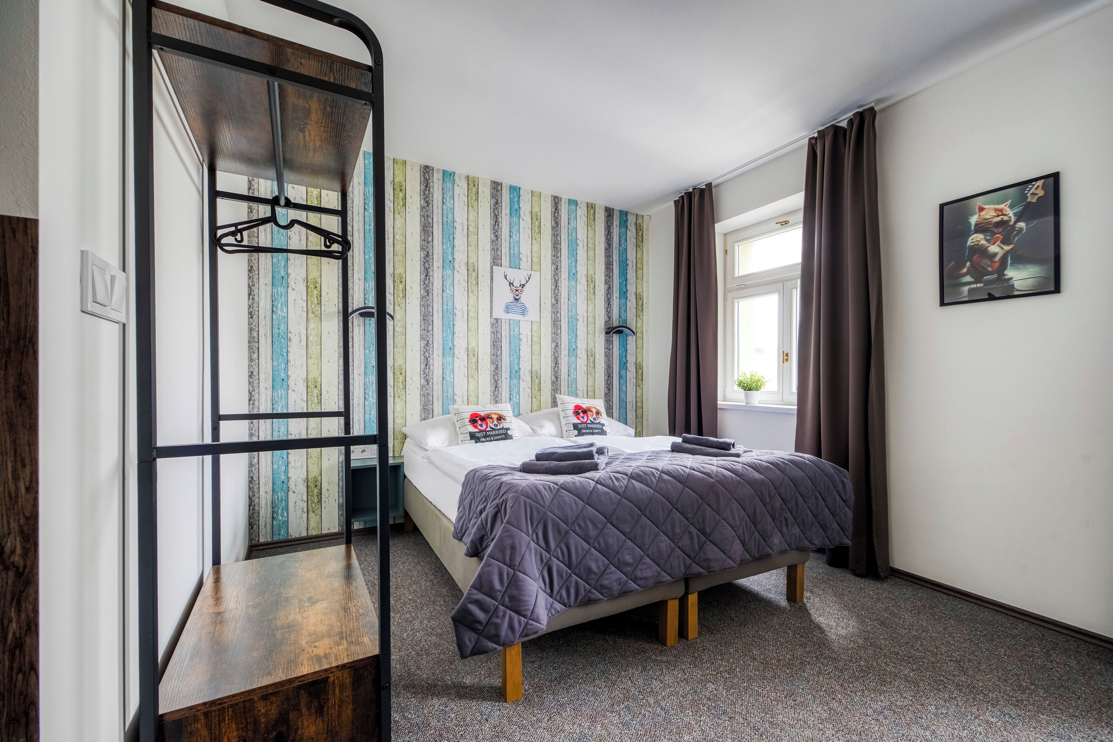 A bedroom with a bed, a hanging coat rack, a striped wall, a window with curtains, and a framed picture on the wall.