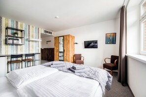 A hotel room with a bed, two chairs, a TV, a shelf with a plant, and a wooden wardrobe.