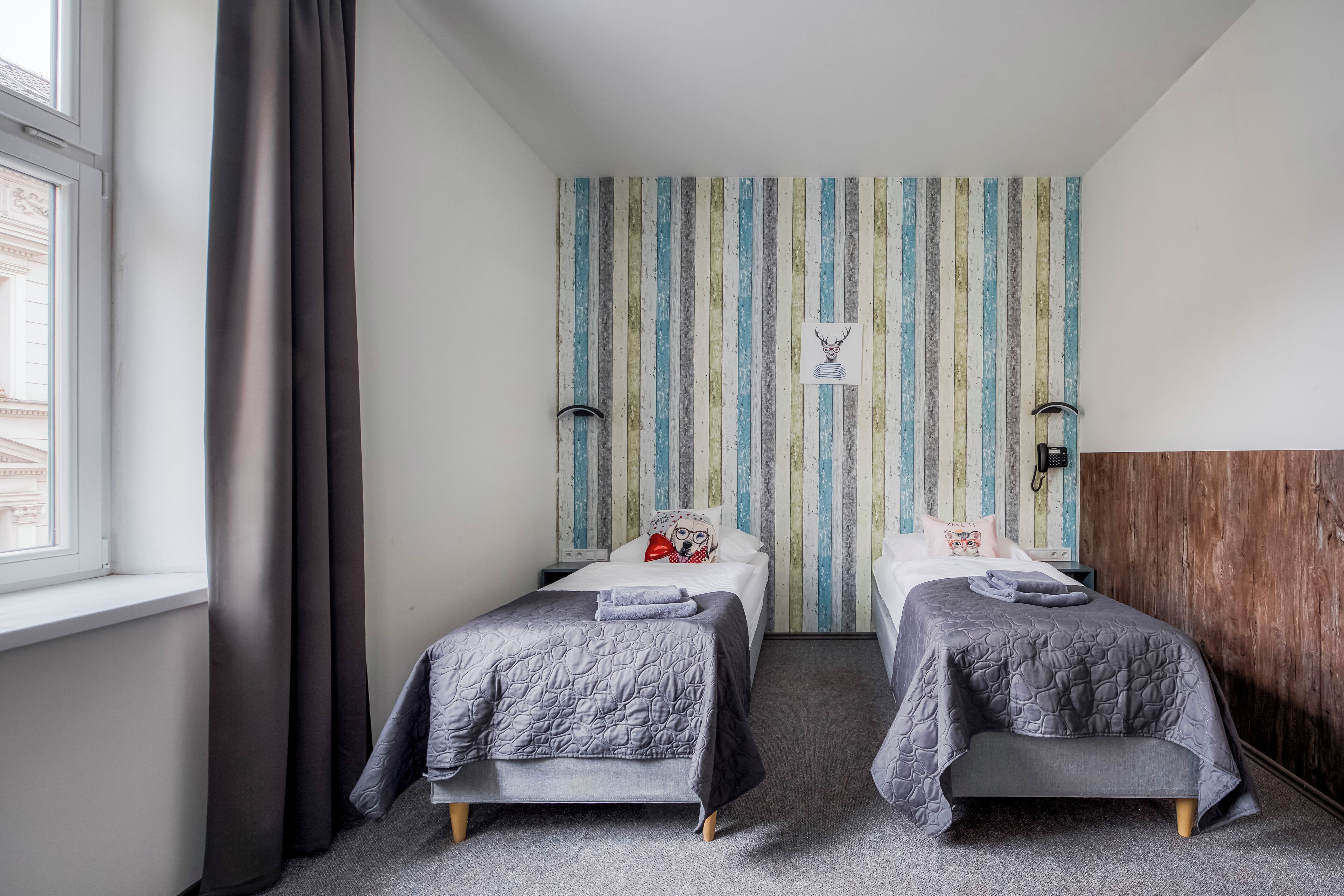 Quarto com duas camas, papel de parede listrado, cabeceira de madeira, janela com cortinas e telefone fixado na parede.
