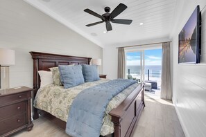 A bedroom with a large bed, a ceiling fan, a nightstand, a lamp, a window with a view of the ocean, and a painting on the wall. - Oceanfront Views! 3 Bed, 2 Bath with Private Beach (Oceanside)