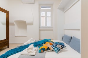A bedroom with a bed, pillows, a blanket, a wooden dresser, a window, and an air conditioning unit.