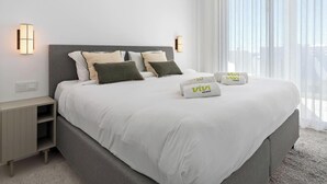 Une chambre d’hôtel moderne avec un grand lit, deux tables de chevet et une vue dégagée à travers des rideaux légers.