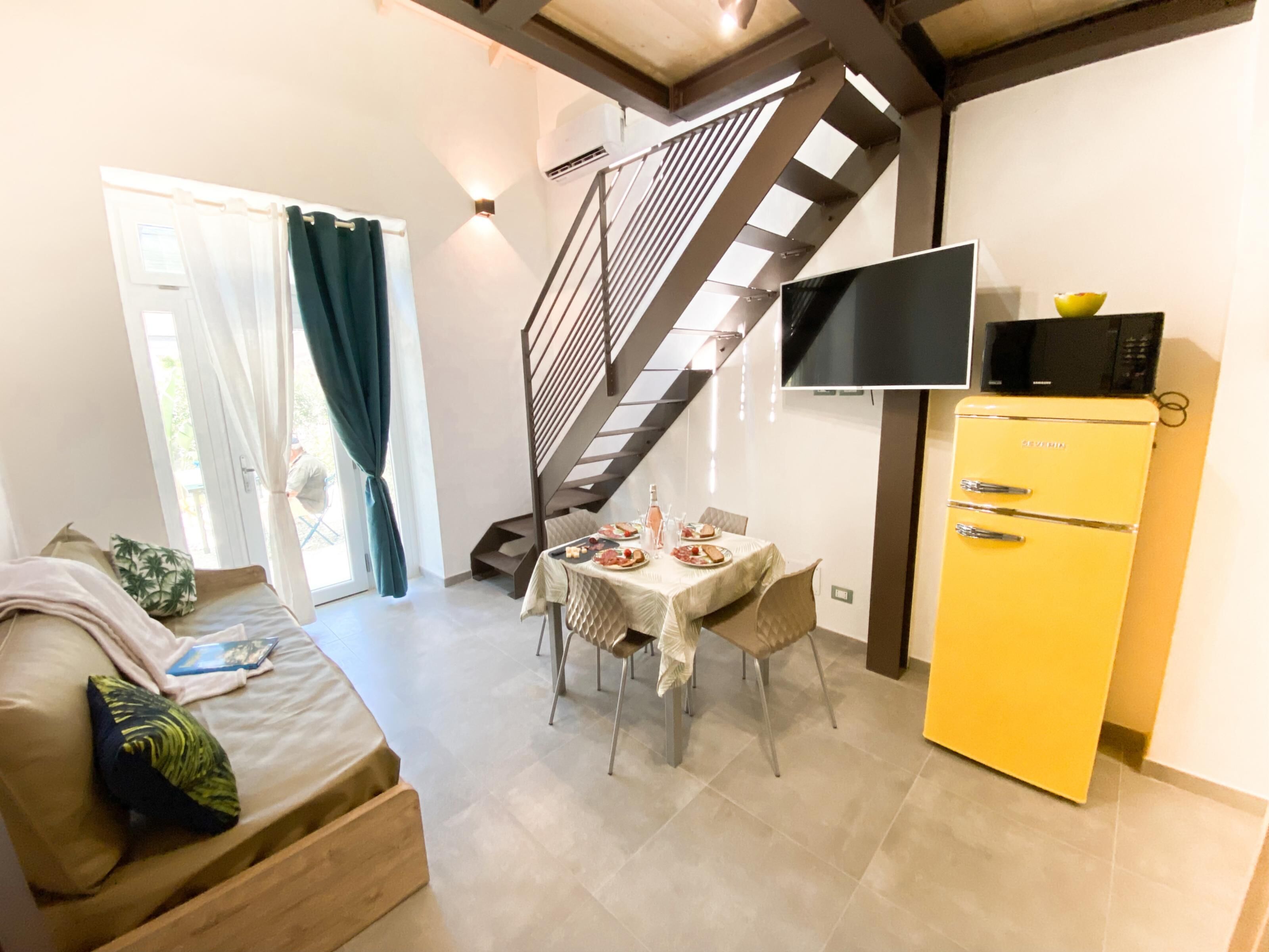 Un espace de vie comprenant un canapé, une table à manger avec des chaises, un réfrigérateur jaune, un téléviseur fixé au mur et un escalier.