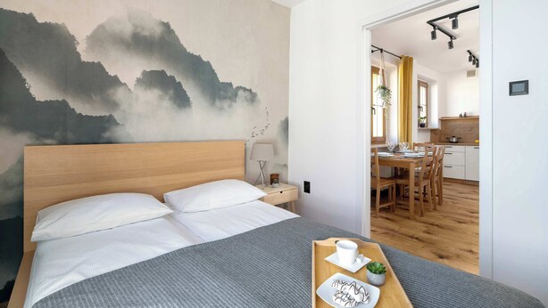 Een slaapkamer met een bed, een houten hoofdbord, een nachtkastje met een lamp en een muur met een schilderij. Een eethoek is zichtbaar door een open deur.