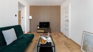 Een moderne woonkamer met een groene bank, een bijzettafeltje met daop een croissant, een tijdschrift en een televisie aan een wand van houten panelen.