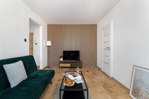 Ein modernes Wohnzimmer mit einem grünen Sofa, einem Couchtisch mit einem Croissant, einer Zeitschrift und einem Fernseher an einer Wand aus Holzpaneelen.