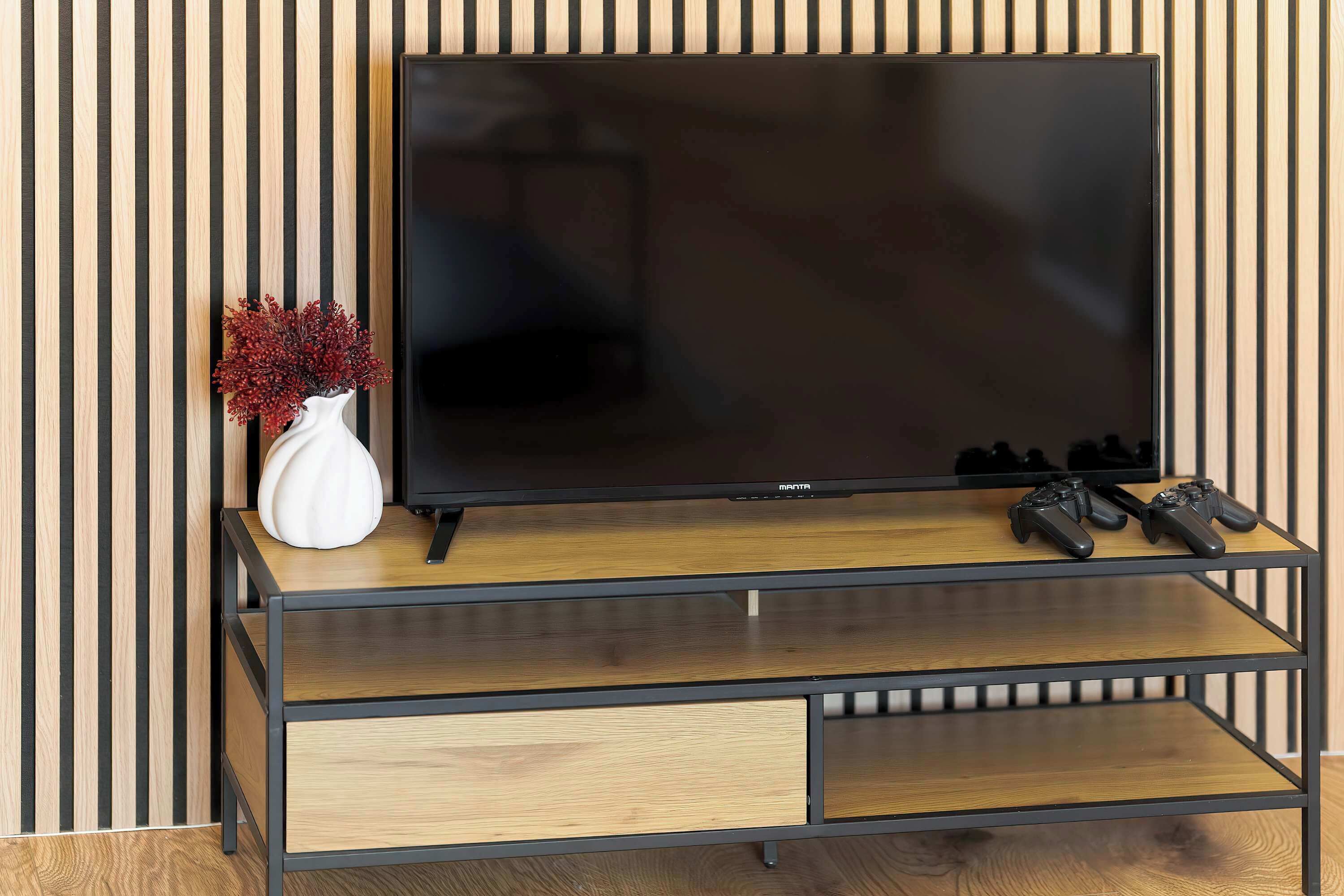 A TV on a wooden TV stand with two game controllers and a white vase with red flowers.