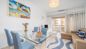 Een moderne woonkamer met een glazen eettafel, blauwe stoelen, een beige bank en een kleurrijk schilderij met een strandthema aan de muur.