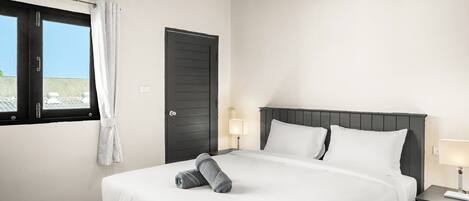 Una cama bien hecha con sábanas blancas y dos toallas enrolladas, una lámpara de noche y una ventana con cortinas.