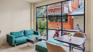 A modern living room with a teal sofa, a wooden coffee table, and a dining table with chairs. A large window offers a view of a red building and a parked scooter outside. - Simply Stay Chaweng (Koh Samui)