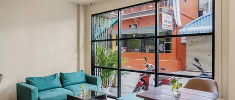 A modern living room with a teal sofa, a wooden coffee table, and a dining table with chairs. A large window offers a view of a red building and a parked scooter outside.