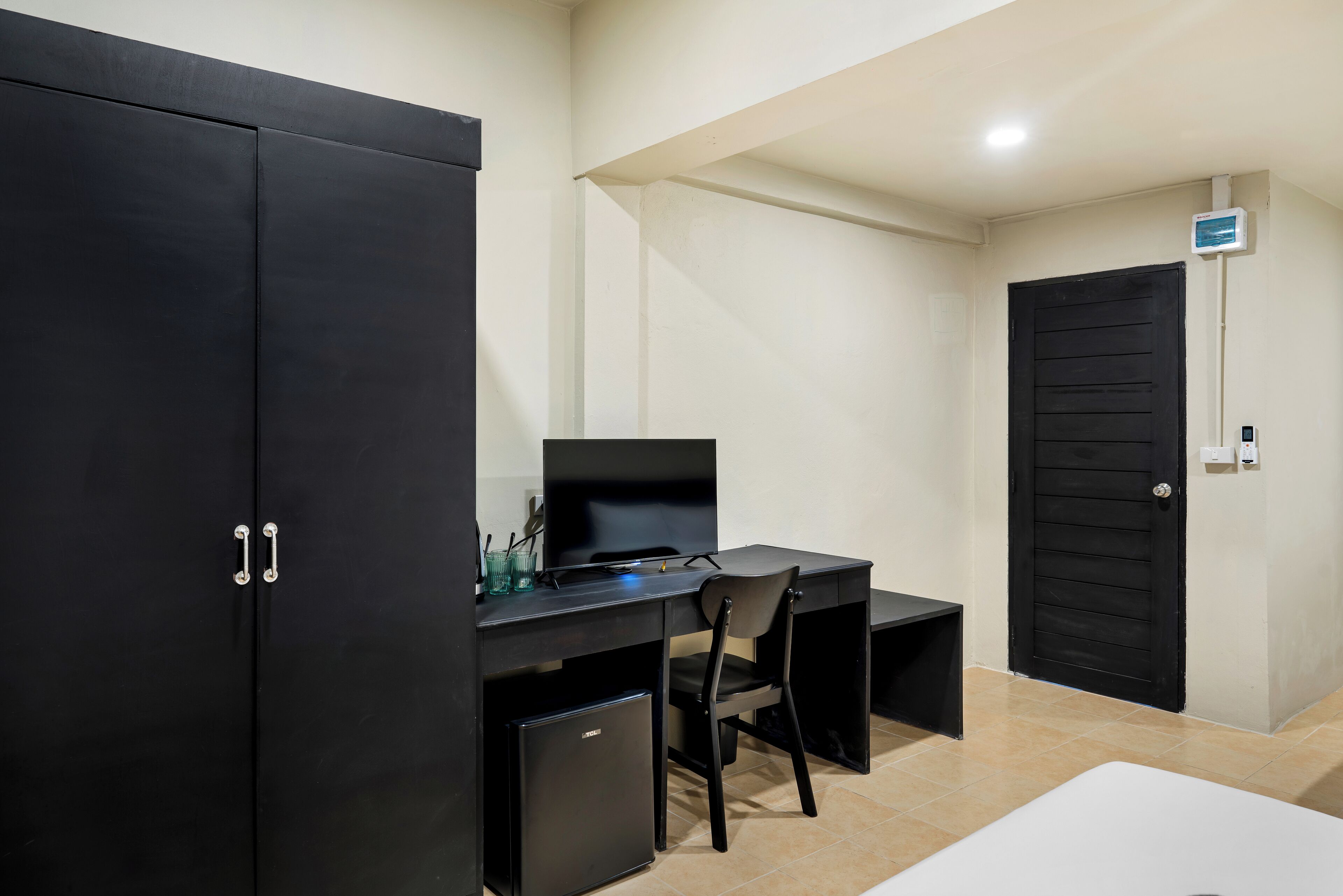 A room with a black wardrobe, a desk with a chair, a television, and a door with a security camera.