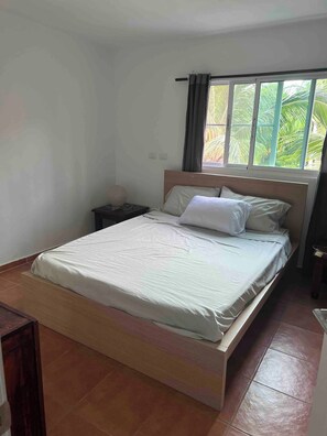 Quarto com uma cama de solteiro, mesa de cabeceira, janela com cortinas e vista para palmeiras.