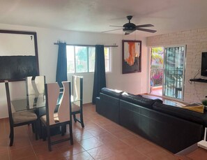 Uma sala de estar com sofá, mesa de jantar, cadeiras, ventilador de teto e uma janela grande com cortinas.