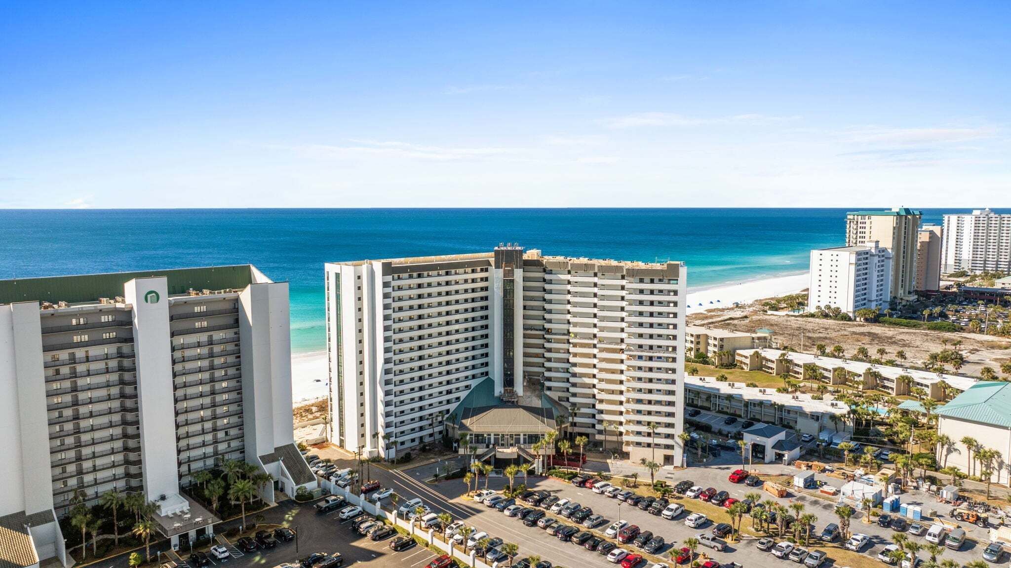 A high-rise hotel with multiple floors, a parking lot, and a beach view.