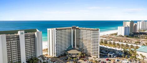 A high-rise hotel with multiple floors, a parking lot, and a beach view.