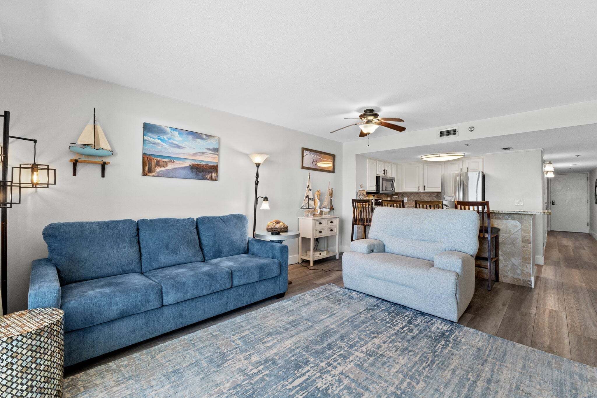 A modern living room with a blue sofa, a light grey armchair, and a patterned rug. A dining area with wooden chairs and a small table is visible in the background.