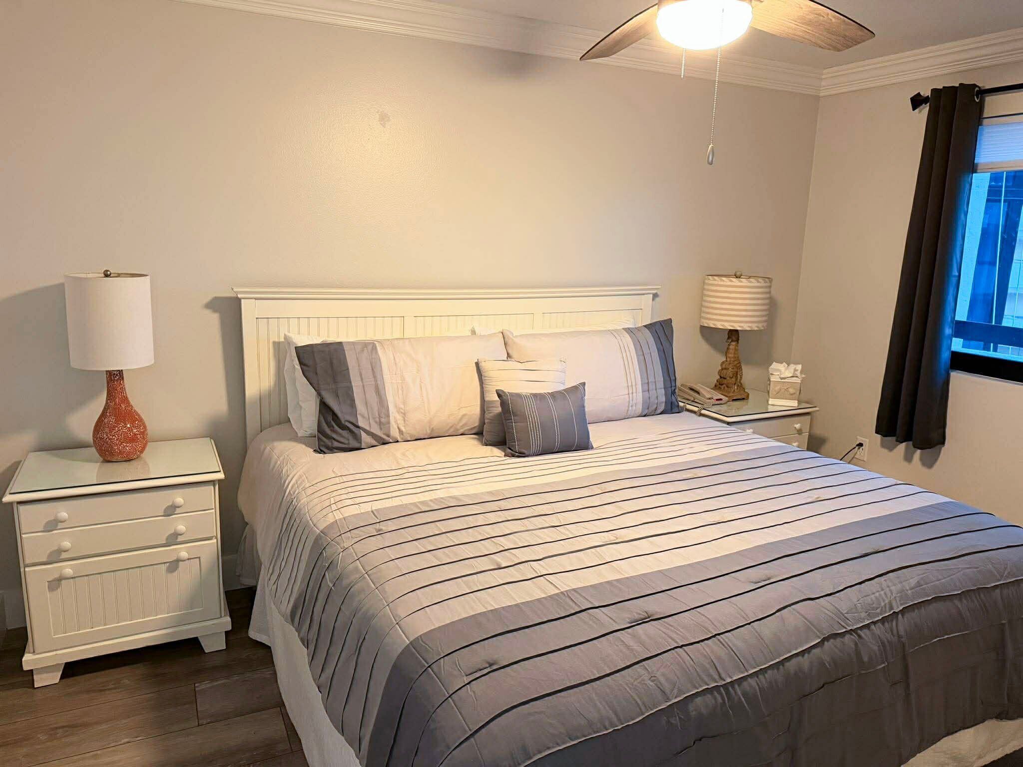 Ein Schlafzimmer mit einem Bett, Nachttischen, einer Lampe, einem Deckenventilator und einem Fenster mit Vorhängen.