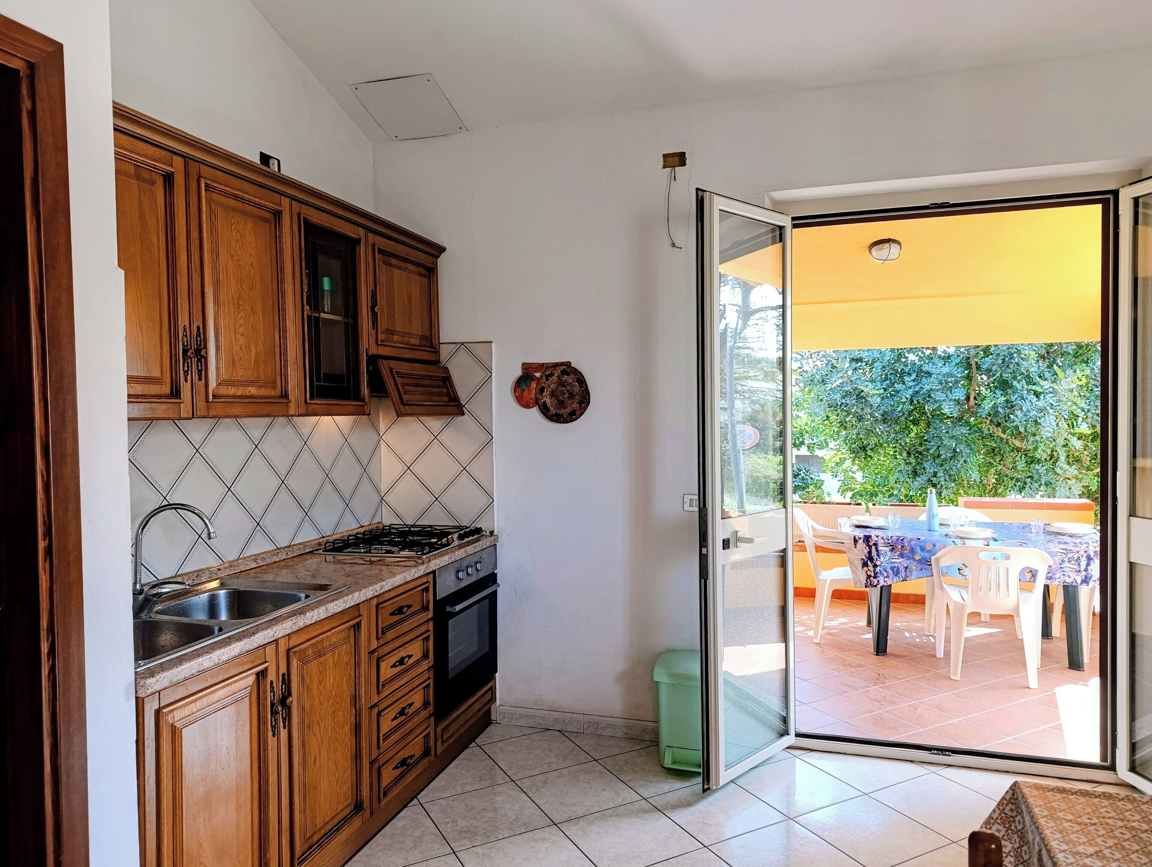 Une cuisine avec des meubles de rangement en bois, une cuisinière à gaz et un évier. Une porte-fenêtre coulissante donne accès à un patio extérieur équipé d’une table et de chaises.