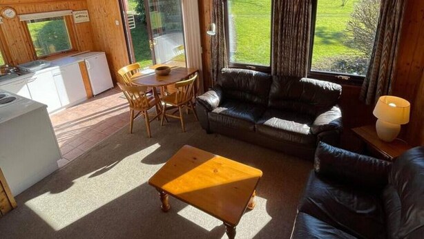 Un acogedor interior de cabaña con techo de madera, un comedor, una sala de estar con sofá y una mesa de centro, y ventanas amplias que ofrecen vistas al exterior.