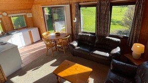 Interior acogedor de una cabaña con techo de madera, zona de comedor, sofá y ventanas amplias con vistas al exterior.