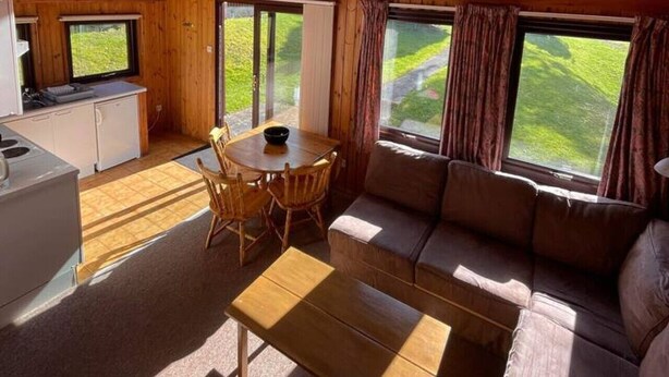Un accogliente interno di una baita con soffitto in legno, zona pranzo con tavolo e sedie, divano e ampie finestre che offrono vista sull'esterno.