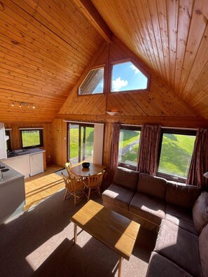 L’intérieur chaleureux d’un chalet, avec un plafond en bois, un coin repas comprenant une table et des chaises, un canapé et de grandes fenêtres donnant sur le paysage extérieur.