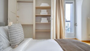 A bedroom with a bed, a closet with folded towels, and a view through a window with curtains.