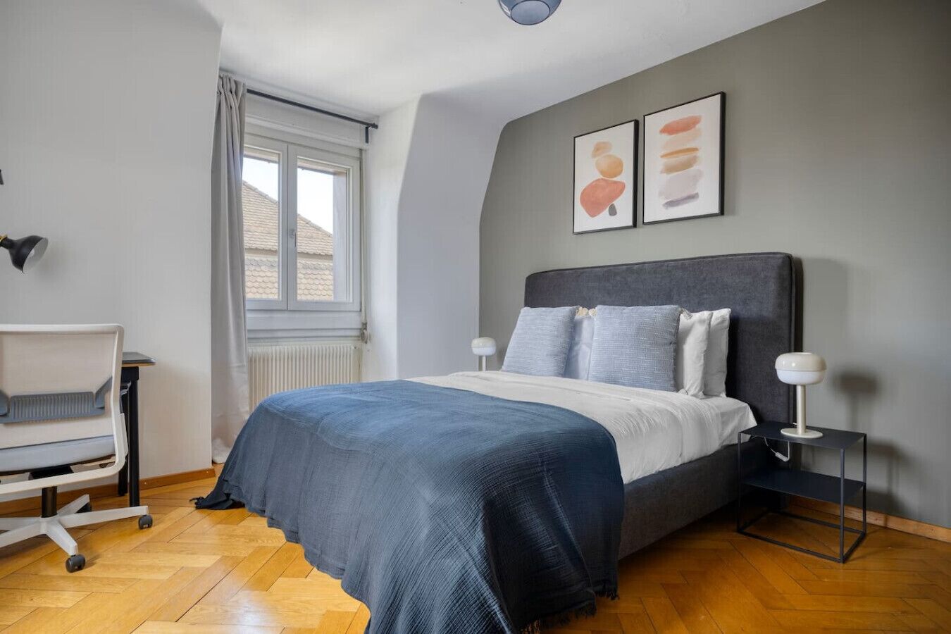 A bedroom with a bed, a desk, and a chair. There is a window with curtains, a radiator, and two wall-mounted lamps.