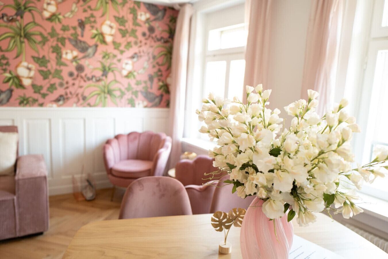 Une pièce avec un papier peint à motifs floraux, un fauteuil rose, une table ornée d’un vase de fleurs et une fenêtre avec des rideaux.