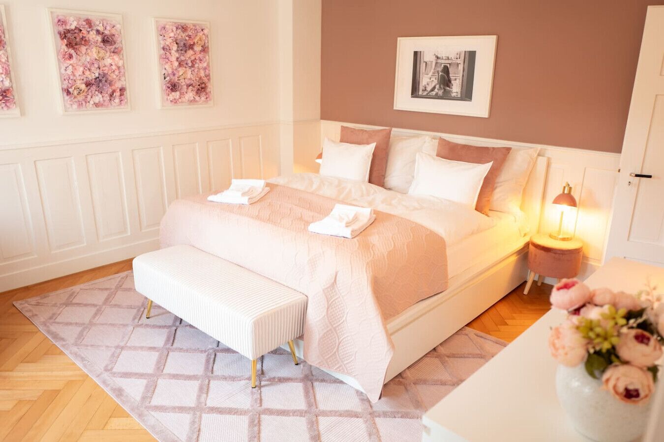 A bedroom with a large bed, a bench, a bedside table with a lamp, and floral decorations.