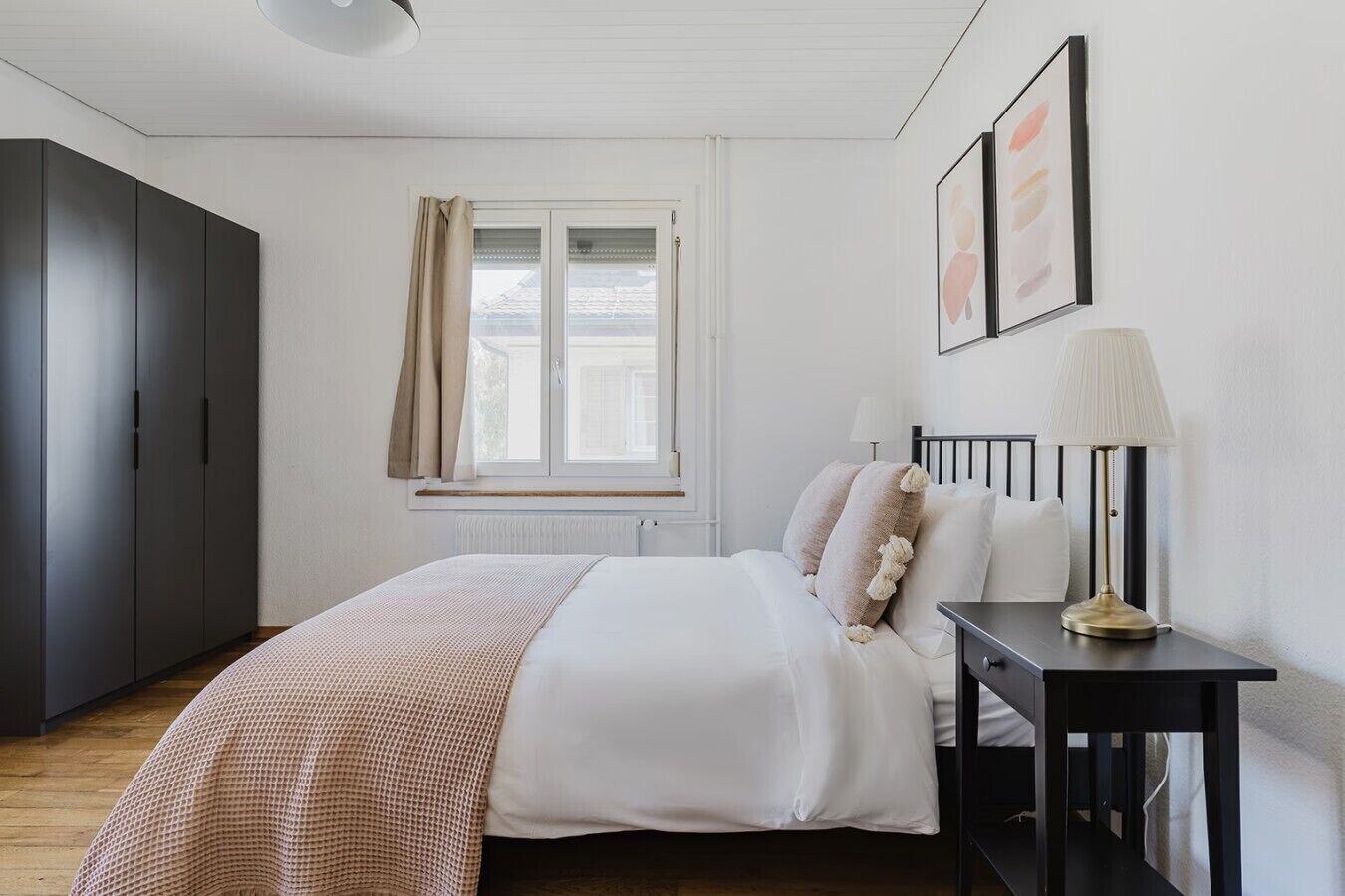 Un dormitorio con cama, mesita de noche, lámpara, armario, ventana con cortinas y una obra de arte en la pared.