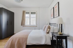 Een slaapkamer met een bed, nachtkastje, lamp, kledingkast, raam met gordijnen en kunst aan de muur.