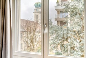 Une fenêtre donnant sur un bâtiment doté d’une tour horloge et sur des arbres sans feuilles, ce qui suggère un décor hivernal.