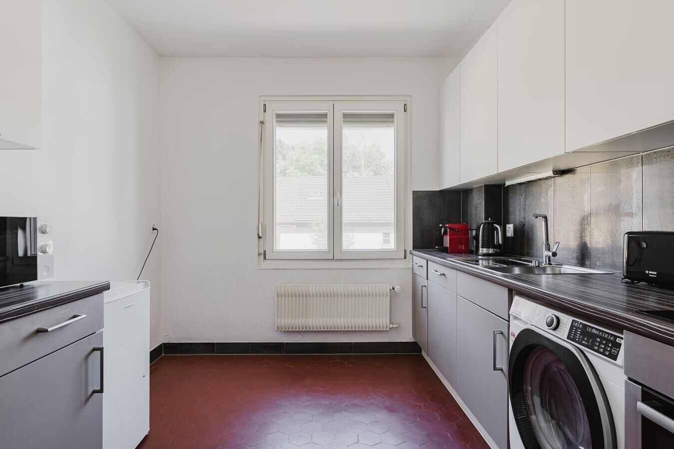 Une cuisine de taille réduite, équipée d’un lave-linge, d’un radiateur et d’une fenêtre avec des stores.