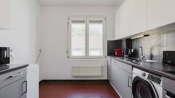 Une cuisine de taille réduite, équipée d’un lave-linge, d’un radiateur et d’une fenêtre avec des stores.