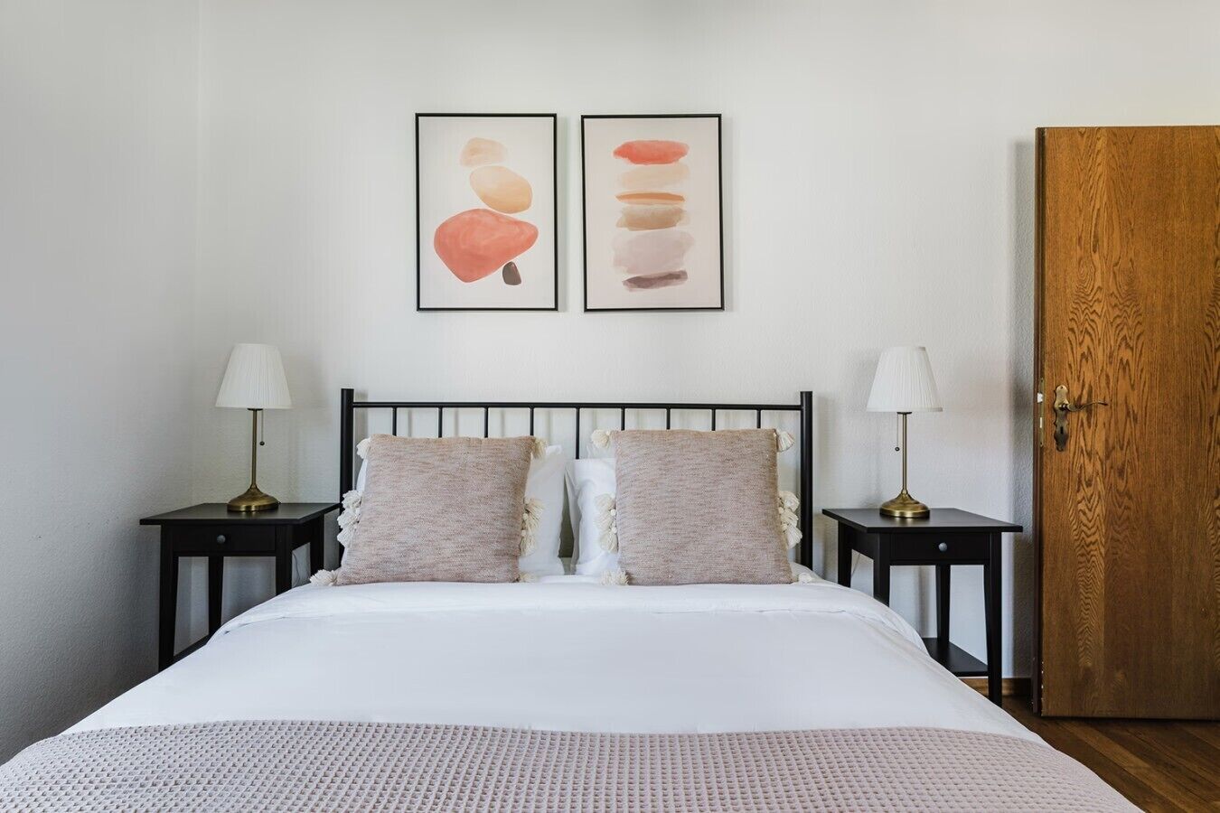 A bedroom with a bed, two bedside tables, two lamps, a wooden door, and two framed artworks on the wall.