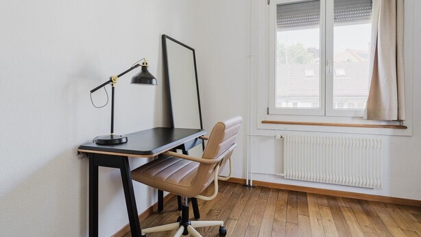 Una habitación con escritorio, silla, lámpara y ventana con cortinas.
