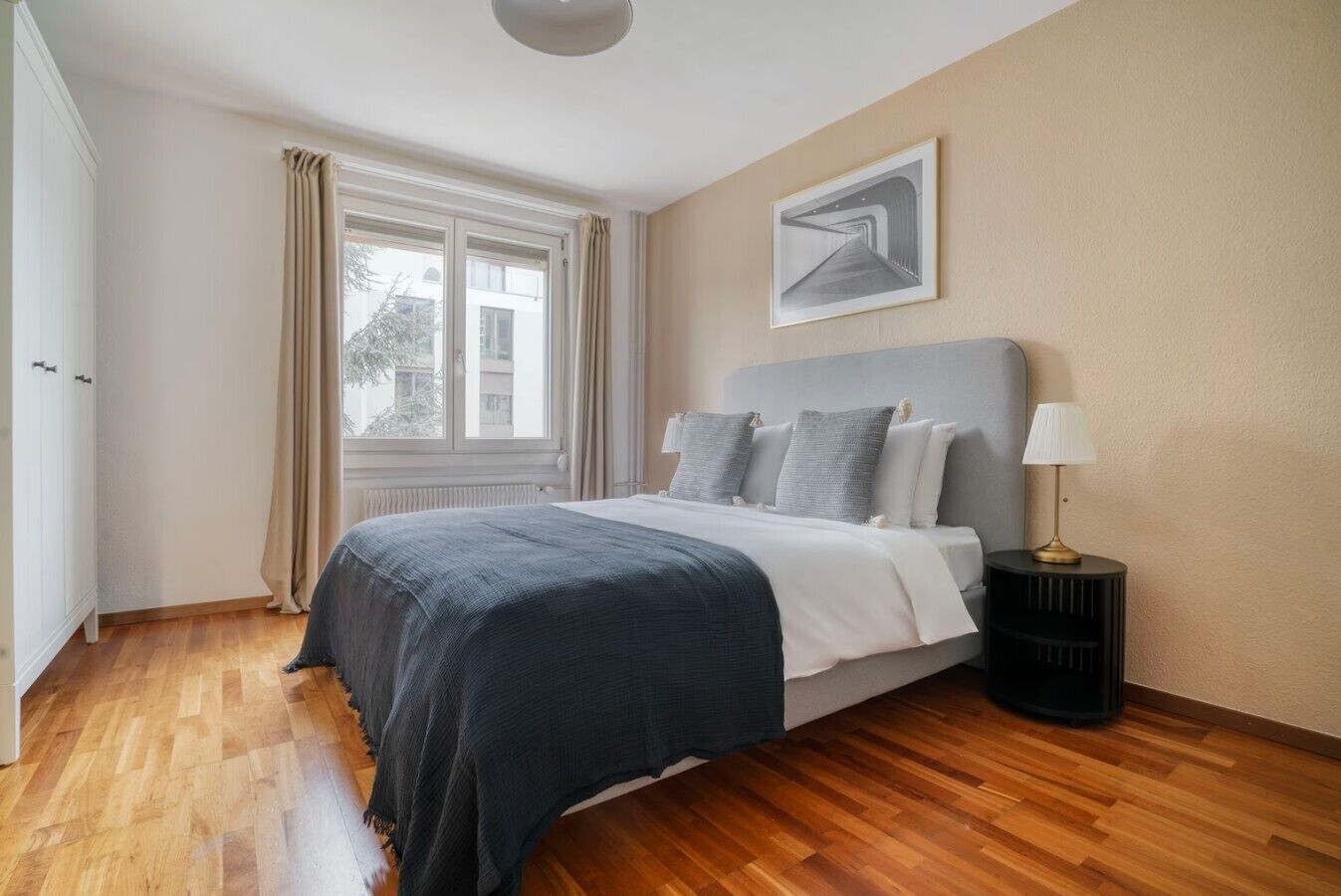 Une chambre à coucher avec un lit, une table de chevet, une lampe, une fenêtre avec des rideaux et un tableau au mur.