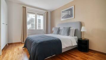 Une chambre à coucher avec un lit, une table de chevet, une lampe, une fenêtre avec des rideaux et un tableau au mur.
