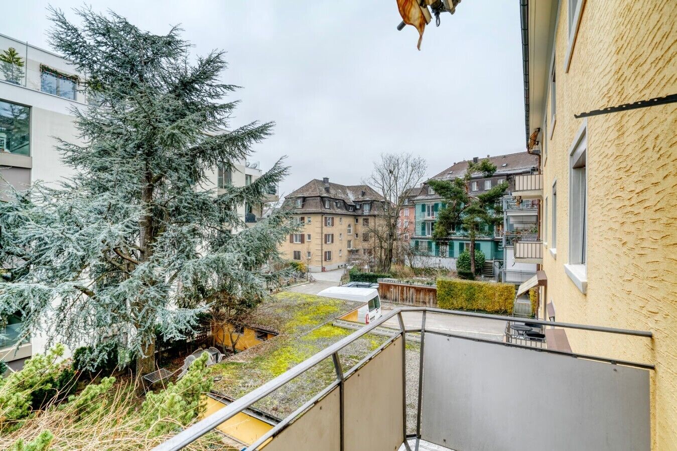 Un quartier résidentiel avec un balcon donnant sur une cour intérieure où se trouve un grand arbre à feuilles persistantes et un petit véhicule blanc.