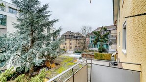 A residential building with a balcony overlooking a courtyard with a large evergreen tree and a small white vehicle.