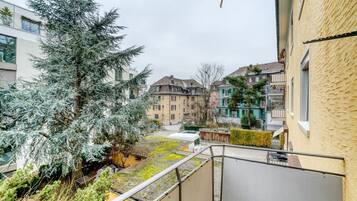 Un quartier résidentiel avec un balcon donnant sur une cour intérieure où se trouve un grand arbre à feuilles persistantes et un petit véhicule blanc.