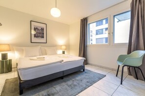 A hotel room with a bed, two bedside lamps, a framed picture on the wall, a window with curtains, and a chair.