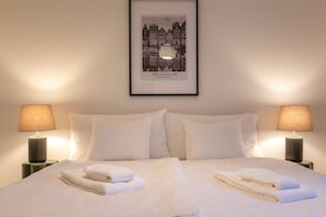 Ein ordentlich bezogenes Bett mit weißen Leinen und Handtüchern, zwei Nachttische mit Lampen und ein gerahmtes Bild von Amsterdam an der Wand.