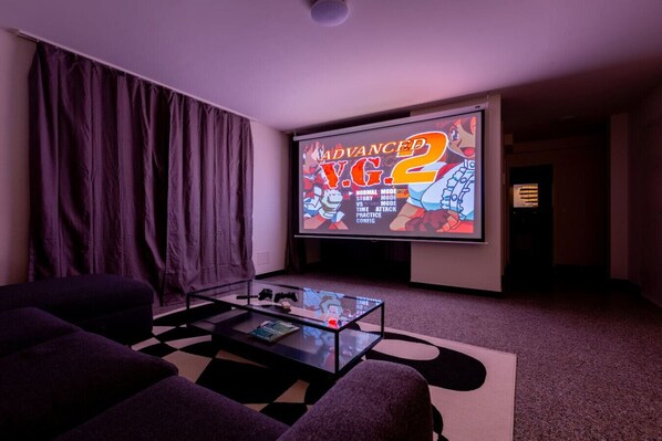 A home theater room with a large screen displaying a video game, a glass coffee table with a game controller, and a dark-colored sofa.